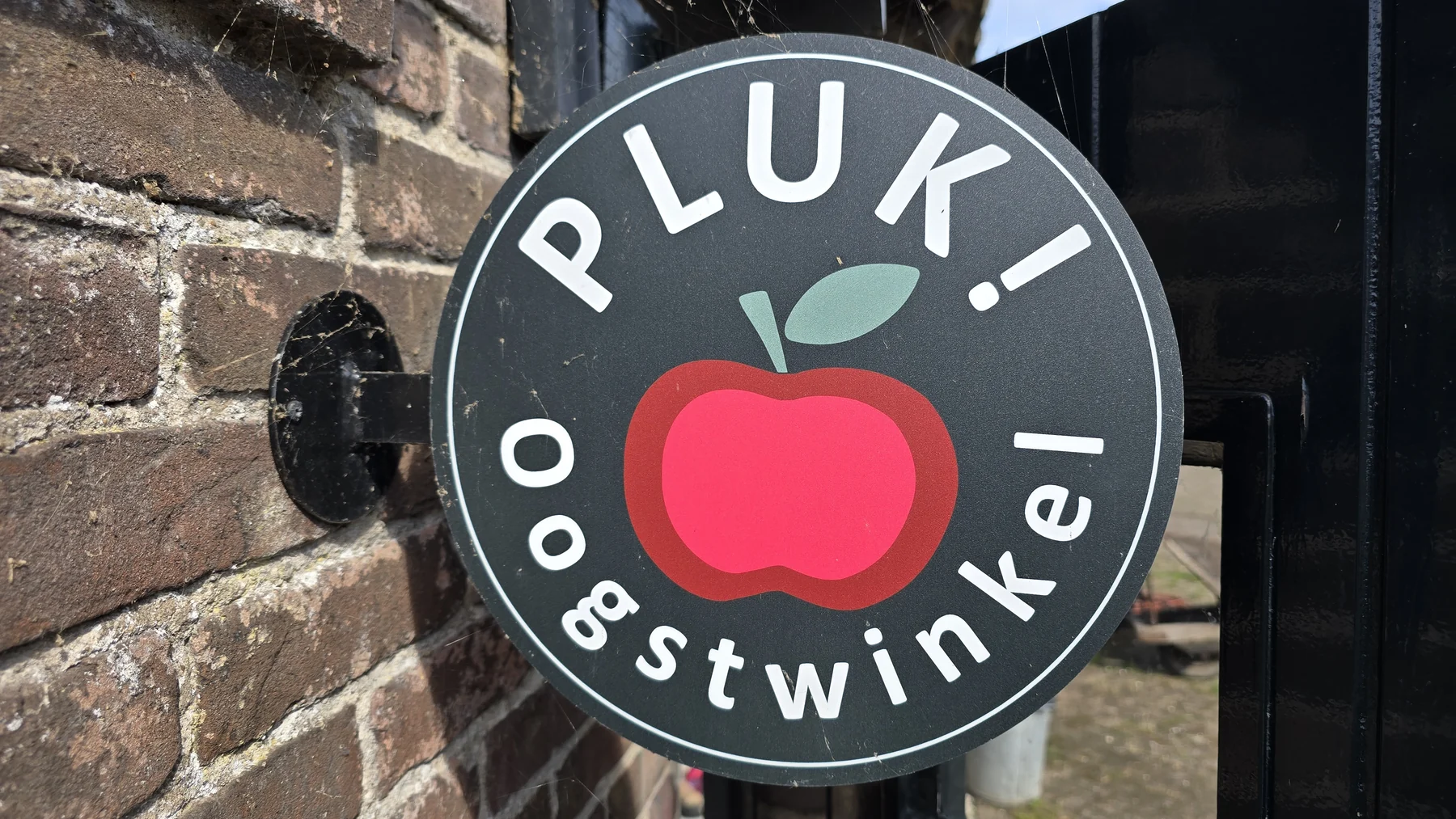Het ronde Pluk!-bord aan de buitenmuur van de oogstwinkel